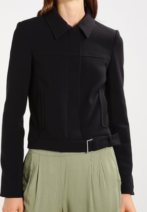 Musta istuva takki, jossa on kaulus, etutaskut ja solkikoriste, yhdistetty korkeavyötäröisiin, vaaleanvihreisiin laskoshousuihin.