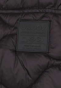 Gevoerde zwarte stoffen vest met een textuur label in zwart met "J&JOY ANDEN LEGACY" en geometrische symbolen, zichtbare gestikte details.