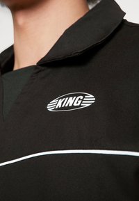 Camicia nera con colletto, caratterizzata da un logo bianco "KING" e dettagli in piping bianco sul petto, realizzata in un tessuto liscio e dalla texture morbida.