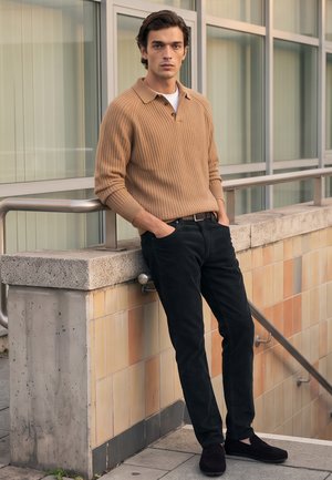 Junger Mann lehnt an Betonwand mit Metallgeländer, trägt braunen Pullover, schwarze Hose und schwarze Schuhe, steht draußen neben den Gebäudefenstern.