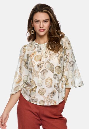 Woman wearing beige blouse with seashell and starfish print and rust-colored pants, posing against plain background.