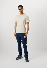 T-shirt beige avec un petit logo noir, associé à un pantalon de sport bleu marine à poches zippées et des chaussures avec des accents gris.
