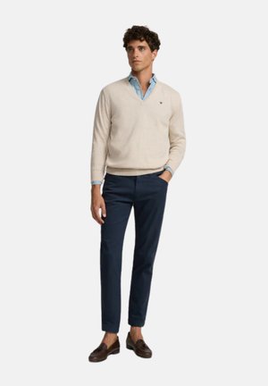 Jersey beige en cuello en V, camisa de color azul claro por debajo, pantalones azul marino, mocasines marrones, postura de pie, cabello corto y rizado, fondo neutro.