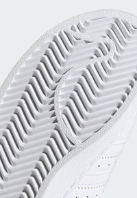 Bijeli potplat tenisice s teksturiranim utorima za prianjanje, s glatkim gumiranim materijalom i blago podignutim rubom. Prostor sa perforiranim uzorkom sa strane.