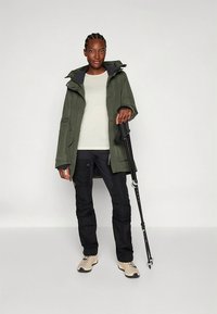 Vodoodporna jakna zelene oljčne barve s kapuco, oblečena čez svetlo srajco. Črne hlače, bež pohodniški čevlji in pohodna palica zaključujejo videz.