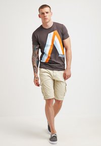 T-shirt marron foncé avec un motif de bandes diagonales bleues et orange, associé à un short cargo beige et des baskets grises.