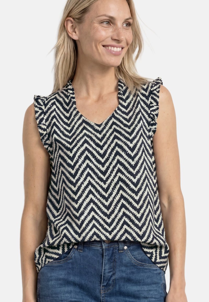 Femme souriante portant un chemisier sans manches bleu marine et blanc à motif chevron, rentré dans un jean bleu sur fond blanc.