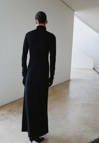 Robe longue noire avec un col haut, un corsage ajusté et des manches longues, associée à des gants noirs. Le tissu semble lisse et élégant.