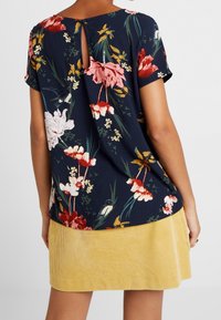 Donna che indossa una blusa a maniche corte floreale blu navy con apertura a chiave sul retro e una gonna in velluto a coste gialla.