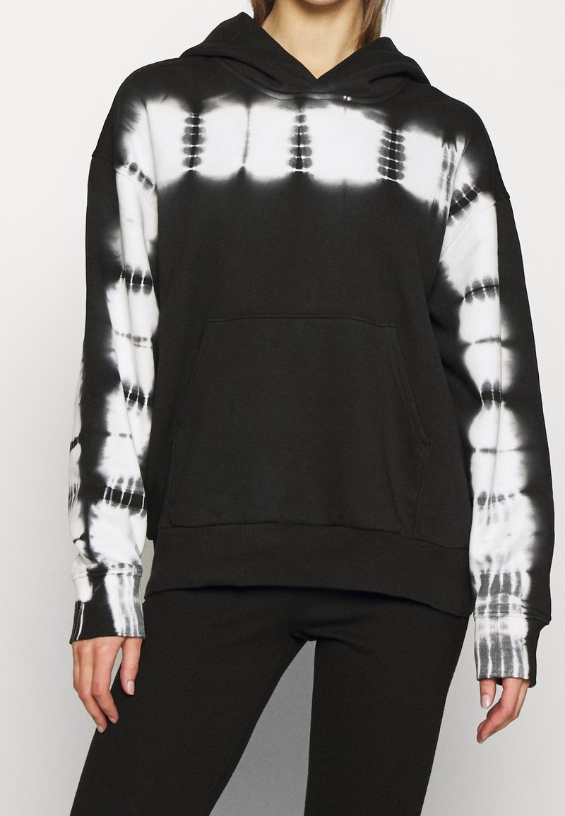 Sweat à capuche noir avec motifs tie-dye blancs sur la poitrine, le haut des bras et les manches, porté avec un pantalon noir sur une personne debout.