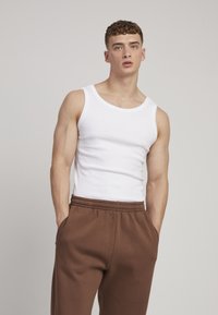 Débardeur blanc à côtes associé à un pantalon de survêtement marron. Le débardeur présente un col rond et un design sans manches. Tissu lisse avec une coupe ajustée.