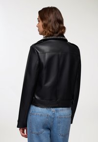 Giacca da biker in pelle sintetica nera con borchie argentate sul colletto, design aderente e taglio cropped, abbinata a jeans in denim azzurro chiaro.