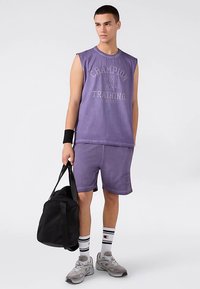 Paarse mouwloze sportshirt met "Champion Training" opdruk, gecombineerd met bijpassende paarse short, zwarte tas en grijze sneakers.