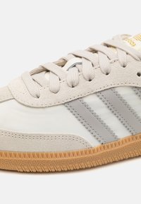 Světlé šedé sneakersy z nubuku a syntetiky se třemi šedými pruhy. Mají kulaté tkaničky a texturovanou gumovou podrážku.