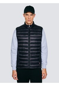 Fiatal férfi fekete steppelt mellényt visel szürke pulóver fölött, és fekete baseballsapkát, világos, egyszerű háttér előtt áll.