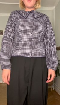 Blusa às quadrados com frente de botão, gola contrastante, mangas longas e punhos franzidos. Usada com calças pretas de cintura alta.