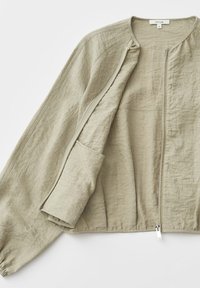 Veste longue en lin beige clair à manches longues avec une fermeture éclair sur le devant, un col rond et des poignets élastiques, posée à plat sur un fond blanc.