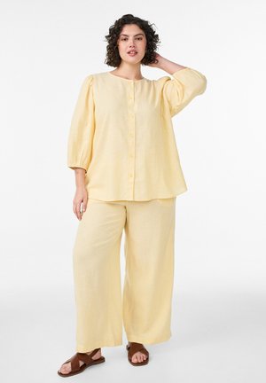 Femme aux cheveux bouclés portant un chemisier ample jaune pâle à boutons et un pantalon à jambes larges, debout avec une main sur la tête et des sandales.