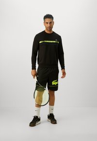 Svart hettegenser med et grønt og lilla logo-stripe, parret med svarte shorts med et grønt krokodille-design. Holder en tennisracket.