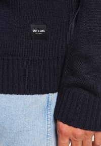 Pull en maille bleu marine avec poignets côtelés, arborant une étiquette carrée noire avec du texte blanc. L'arrière-plan montre un tissu en denim bleu clair.