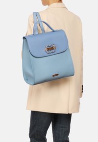 Zaino in pelle blu con pattina, hardware dorato, dettagli intrecciati e doppio manico. Indossato a tracolla, abbinato a un blazer neutro.