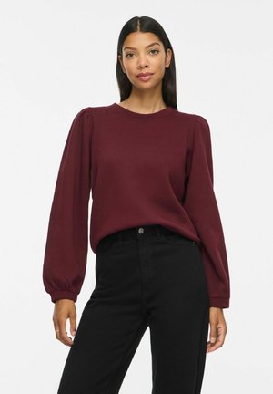 Femme aux cheveux raides et foncés portant un haut bordeaux à manches longues et un pantalon noir taille haute, debout devant un fond uni.