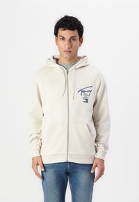 Svijetlosmeđi zip-up dukserica s prednjim džepom, kapuljačom s uže i mornarsko plavim logom. Uparen s plavim trapericama.