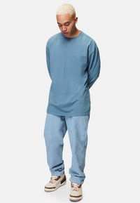 Långärmad, ljusblå sweatshirt med en broderad logotyp, kombinerad med löst sittande ljus denimjeans och beigefärgade träningssneakers.