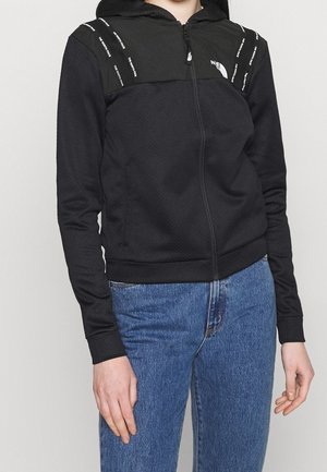 Personne portant un sweat à capuche zippé noir avec le logo "The North Face" et un jean bleu, visible du cou aux hanches sur fond uni.