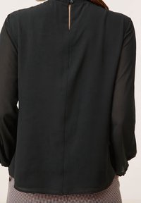 Femme portant un chemisier noir à manches longues avec une ouverture en forme de goutte d'eau et un bouton dans le dos, associé à un pantalon à motifs.