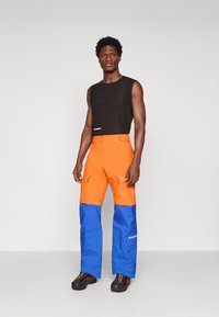 Ανδρας φορά μαύρο αμάνικο πουκάμισο, πορτοκαλί και μπλε παντελόνια εξωτερικού χώρου και μαύρες ορειβατικές μπότες, στέκεται μπροστά από ένα απλό ανοιχτό φόντο.