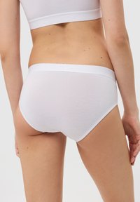 Frau in weißen Baumwollunterhosen und einem passenden, kurzen Tanktop, von hinten vor einem schlichten Hintergrund gezeigt.