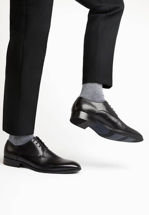 Man in zwarte nette schoenen en grijze sokken met één voet opgetild, gekleed in zwarte formele broek tegen een witte achtergrond.