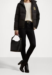 Nainen, jolla on päällään musta untuvatakki, vaalea palmikkoneulepaita, mustat kapeat farkut, musta vyö, mustat korkeat saappaat ja h än pitää mustaa nahkakäsilaukkua.