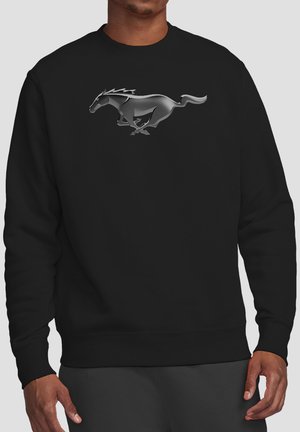 Man wearing black sweatshirt with shiny silver running horse graphic centered on the chest against a plain light background.
