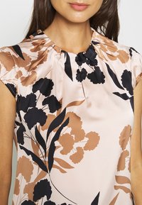 Bluse mit Blumenmuster, die einen sanften rosa Hintergrund, schwarze und braune Blumenmuster, eine gefaltete Halslinie und kurze Ärmel aus satinartigem Stoff aufweist.