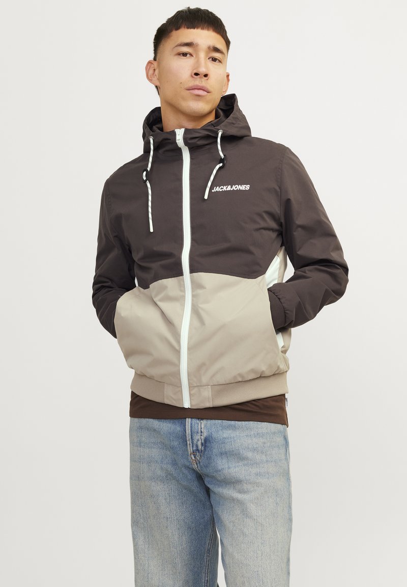 Lightweight, two-tone jacket in dark brown and beige, featuring a zip front, hood, white accents, and a logo on the chest.