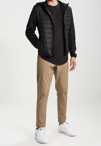 Mężczyzna ubrany w czarną pikowaną kamizelkę, czarną bluzę z długim rękawem, beżowe spodnie i białe trampki, stojący z jedną ręką w kieszeni.