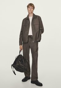 Giacca brown con zip e due tasche sul petto, abbinata a pantaloni coordinati. Scarpe in pelle nera e uno zaino scuro con trama.
