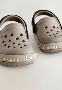 Beżowe klapki z otworami wentylacyjnymi, czarnym paskiem i teksturowaną powierzchnią. Na boku wytłoczone białe logo "Ted Baker". Gumowa podeszwa.