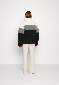 Giacca imbottita con colletto alto, presenta strisce orizzontali in nero, grigio e bianco, realizzata in materiale peluche. Indossata sopra pantaloni color crema.