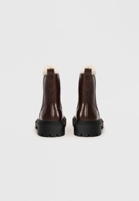 Tmavě hnědé kožené kotníkové boty s černými robustními podrážkami a bílým kožíškem viditelným na vrchu, zobrazené zezadu na jednobarevném pozadí.