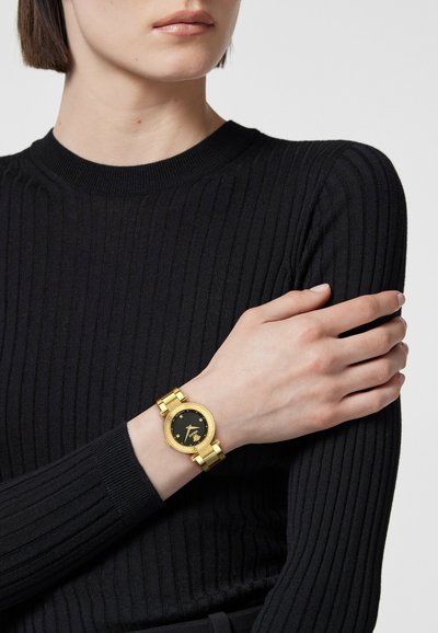 Guldmetal ur med en rund sort urskive og guldkant, der har en kædet rem. Båret på et bar håndled mod en sort ribbet sweater.