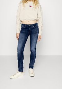 Sudadera corta beige con cintura ajustable de cordón, jeans ajustados azul oscuro y zapatillas blancas con acentos multicolores. Textura de tela suave.