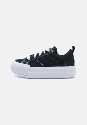 Czarny niski sneaker z grubą białą gumową podeszwą, białym przeszyciem, białymi sznurowadłami i pętelką na pięcie, na jednolitym jasnym tle.