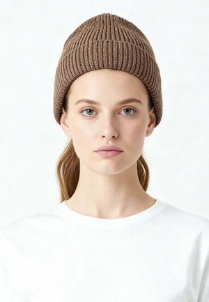 Jeune femme à la peau claire, aux yeux verts et aux cheveux châtain clair, portant un bonnet en tricot marron et un haut blanc, faisant face à l'avant sur un fond uni.