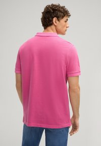 Roze katoenen polo met een klassieke kraag, korte mouwen en een rechte zoom. Model wordt van achteren weergegeven met blauwe jeans.