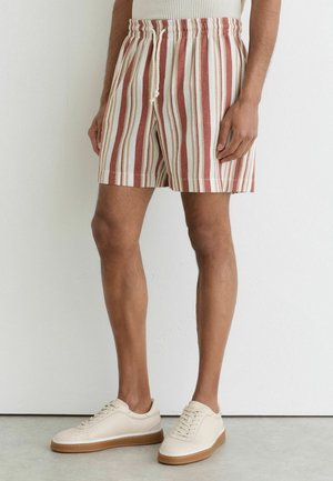Mand iført knælange shorts med lodrette røde og hvide striber og beige sneakers med gummisåler står op ad en hvid væg.