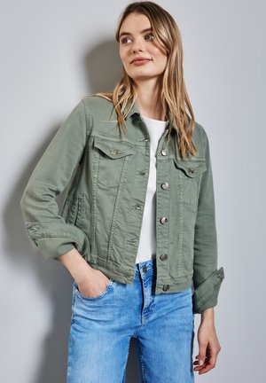 Grön denimjacka med knapplist, två bröstfickor och uppvikta ärmar, tillsammans med ljusblå jeans och en vit t-shirt under.