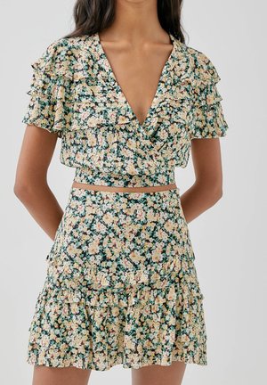 Kvinde iført en blomsterprintet, flæset crop top med matchende højtaljet nederdel, med gule og hvide blomster på en mørkegrøn baggrund.